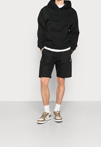 Černá mikina s kapucí a přední kapsou, doplněná černými cargo šortkami s bočními kapsami. Na nohou bílé ponožky a hnědé tenisky.