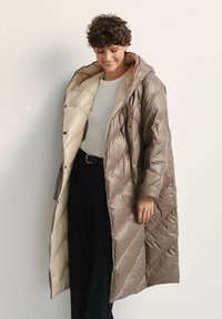 Długi puchowy płaszcz w odcieniach beżu i jasnego brązu, z diamentowym wzorem pikowania, kapturem oraz zapięciem na zatrzaski. Miękka, błyszcząca faktura.