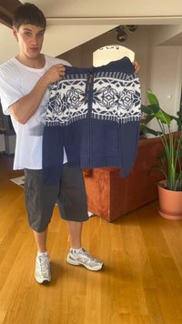 Giovane in t-shirt bianca e pantaloni corti che tiene un maglione con zip di colore blu navy e bianco a fantasia in un soggiorno con pavimento in legno.
