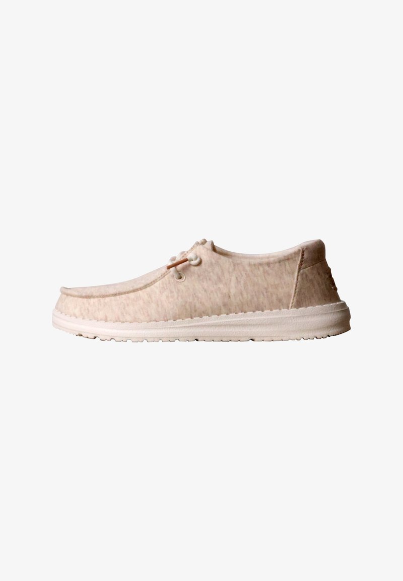 Chaussure décontractée beige sans lacets avec dessus en tissu doux, semelle en caoutchouc blanche et détail de lacets latéraux sur fond blanc.
