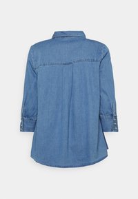 Camicia di denim con yoke posteriore, maniche lunghe e polsini con bottoni a scatto. Colore azzurro chiaro, texture morbida, orlo curvo e dettagli di cucitura sottili.