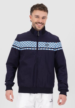 Marinefarbene Windjacke mit Stehkragen, die ein hellblaues kariertes Muster über der Brust aufweist und elastische Bündchen hat. Logo auf der Brust.