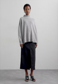 Un pull surdimensionné gris clair avec un col montant, associé à une jupe noire longueur cheville avec une fente avant. Des talons ouverts noirs.