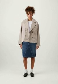 Woman with curly hair wearing beige double-breasted jacket, white top, denim skirt, white socks, and black loafers, standing against plain background.