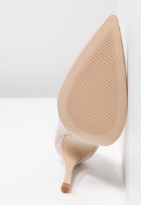 Nude högklackad sko med spetsig tå och en elegant design. Sålen är texturerad med horisontella spår för grepp.