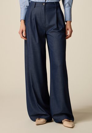 Donna che indossa pantaloni a vita alta, a gamba larga di colore blu scuro abbinati a ballerine beige con punta appuntita e dettaglio catena, in piedi davanti a uno sfondo semplice.