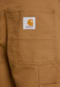 Close-up of tan fabric pocket with white rectangular Carhartt brand label sewn near the top center.
