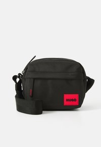 Kompakte schwarze Umhängetasche mit verstellbarem Riemen, Front-Reißverschluss und rotem Hugo-Logo-Patch in der unteren rechten Ecke.