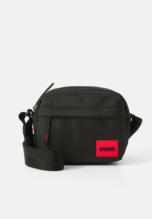 Geantă crossbody compactă, de culoare neagră, cu curea ajustabilă, fermoar frontal și patch cu logo roșu Hugo în colțul din dreapta jos.