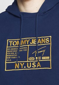 Tmavě modrá mikina s kapucí, zdobená velkým žlutým vyšívaným motivem s textem a designem globu, vyrobená z měkké bavlny s kapucí na stahovací šňůrku.