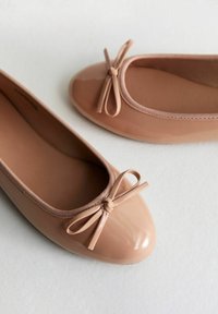Nude ballet flats with a glossy finish, round toe, and a bow accent. Made of flexible material with a smooth texture and cushioned interior.