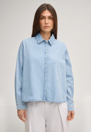 Frau mit glattem braunem Haar, die ein hellblaues Jeanshemd mit Knöpfen und weiße Hose trägt, steht vor einem schlichten hellen Hintergrund.