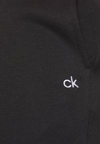 Svart bomullstyg med en slät yta, försett med en vit broderad "ck"-logotyp nära halsringningen. Klassisk design.