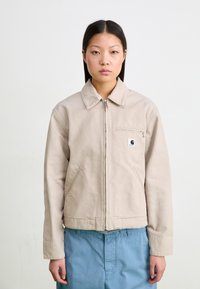 Beige lukuga, millel on pehme tekstuur, suur krae, rinna tasku ja küljetaskud; kantud helesiniste pükste peal.