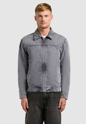 Muž s krátkymi blond vlasmi, oblečený v vybledlej sivej bunde na zips a tmavých džínsoch, stojaci pred jednoduchým svetlým pozadím.