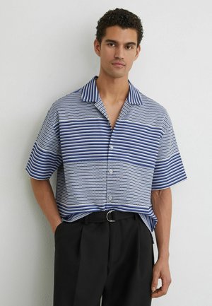 Férfi rövid ujjú, kék-fehér csíkos gombos ingben és fekete nadrágban, egy sima fehér fal előtt állva.