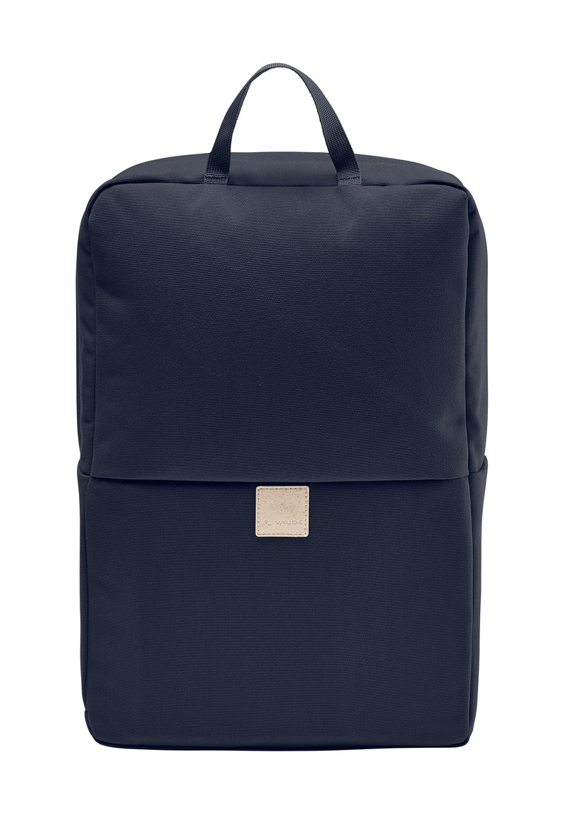 Mochila de tela azul marino con forma cuadrada, asa superior y un parche de logo beige. Textura suave con diseño minimalista.