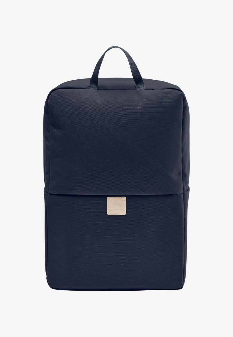 Navy blue fabric backpack with a square shape, top handle, and a beige logo patch. Smooth texture with minimalistic design.