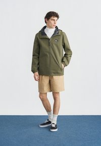 Νεαρός άνδρας που φοράει χακί ζακέτα με φερμουάρ, μπεζ σορτς, άσπρες κάλτσες και μαύρα αθλητικά παπούτσια, στέκεται σε μπλε δάπεδο μπροστά σε άσπρο τοίχο.