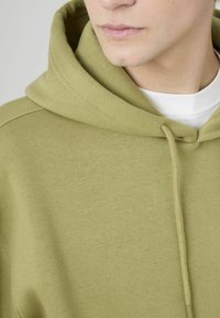 Sweat à capuche vert olive en mélange de coton doux. Comprend une capuche avec cordon de serrage, un tissu texturé et un col en côtes, montrant une vue rapprochée.