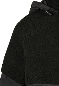 Svart fleece yttertøy med en teksturert overflate, utstyrt med hette i nylon og lukkeanordninger, designet for varme og komfort.