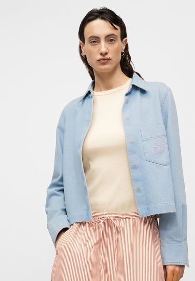 Ανοιχτό γαλάζιο τζιν μπουφάν με μια τσέπη που έχει ροζ κέντημα, φορεμένο πάνω από μπεζ τοπ και πορτοκαλί παντελόνια με λωρίδες και ελαστική μέση.