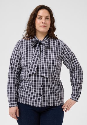 Vrouw met krullend bruin haar die een zwart-wit geruit overhemd met knopen en vlinderdetail draagt, staand met één hand op haar heup.
