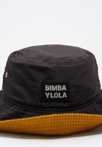 Sombrero de cubo negro con ojales dorados y un ala texturizada en mostaza, que presenta un parche negro con texto blanco "BIMBA Y LOLA".