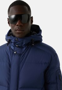 Marineblå pufferjakke med høj krave og justerbar hætte, med trykknaplukninger og en lynlåslomme i siden. Sorte solbriller.