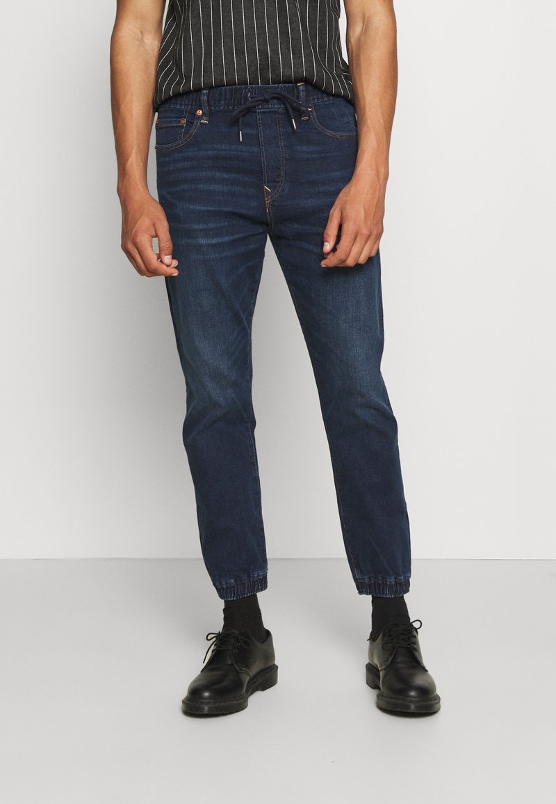 Homme portant un jean jogger bleu foncé avec des poignets élastiques, des chaussures habillées noires et un débardeur rayé noir et blanc devant un mur uni.