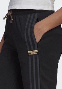 Hand i fickan på svarta Adidas-byxor med tre mörka ränder och beiga Adidas-logotypen, i kombination med vit skjorta.