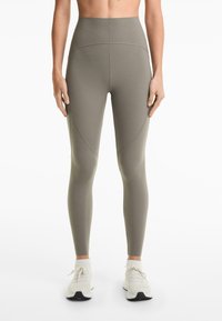 Leggings taille haute dans un vert olive atténué, avec un tissu lisse et extensible et des détails de coutures subtils ; associés à des chaussures de sport blanches.