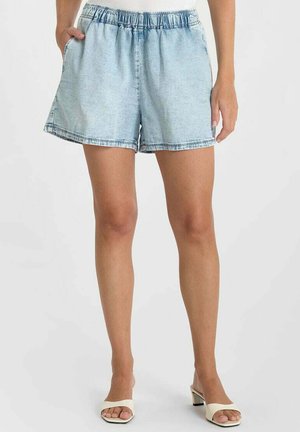 Femme portant un short en denim bleu clair à taille élastique avec poches latérales et des sandales blanches à talon bloc ouvertes à l'avant, debout devant un fond blanc.