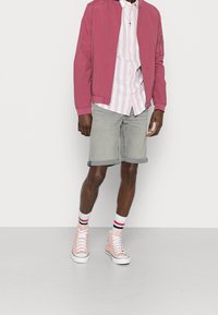 Giacca bomber rosa chiaro sopra una camicia a righe rosa e bianca, pantaloni corti grigi arrotolati e sneaker alte rosa con suole bianche e calze a righe.
