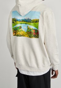 Hellgrauer Kapuzenpullover mit einem großen bunten Landschaftsprint auf der Rückseite, der einen See, Berge und lebendige Blumen zeigt.