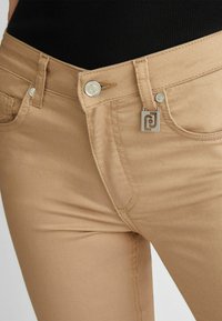 Jeans beige con una texture liscia, dotati di un bottone argentato, un piccolo marchio e cuciture dettagliate lungo le tasche.