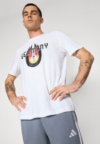 Hvid bomulds T-shirt med et sort og rødt grafik, der har ordet "Tyskland" og et emblem i gul og sort. Standard rund hals.
