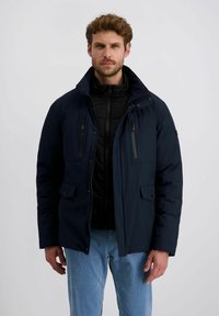 Mann in einer marineblauen Parkajacke über einem schwarzen Reißverschluss-Pullover und hellblauen Hosen, der vor einem schlichten weißen Hintergrund steht.