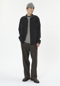 Sort skjorte med knapper, stribet grå og hvid t-shirt under, brune bukser, grå strikket beanie og mørke sko. Enkel, struktureret design.