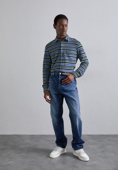 Μαύρο και πράσινο πουκάμισο με ζιγκ-ζαγκ σχέδιο, συνδυασμένο με μπλε τζιν και λευκά αθλητικά παπούτσια, το οποίο στέκεται μπροστά από μια ουδέτερη διακόσμηση.