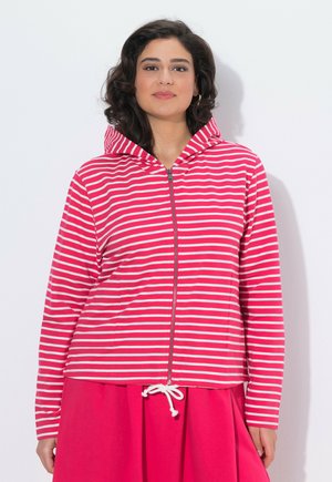 Ung kvinde iført en rød og hvidstribet hættetrøje med lynlås og hætte på, kombineret med et ensfarvet rødt nederdel med hvid snor.
