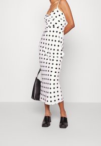Kvinna klädd i en vit midi-slipklänning med svarta prickar, svarta muleklackar och bärande en svart handväska mot en enfärgad bakgrund.