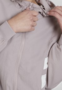 Hænder der justerer snoren og lynlåsen på en lys mauve vindjakke med ribbede manchetter og hvide siderændebånd.