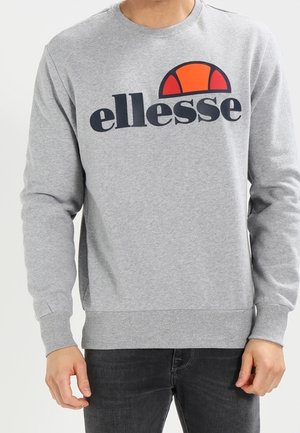 Mann trägt grauen Sweatshirt mit schwarzem und orangefarbenem „ellesse“-Logo auf der Brust, kombiniert mit dunklen Jeans.