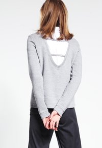 Osoba ubrana w jasnoszary top z długim rękawem z głębokim wycięciem na plecach, nosząca biały podkoszulek, z rękami złożonymi za plecami.