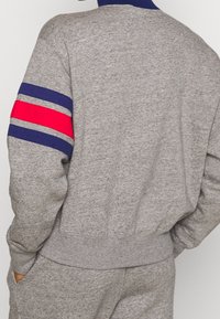 Sudadera gris con cuello y puños acanalados. Presenta rayas horizontales en azul marino y rojo en la manga izquierda. Tejido suave y texturizado.