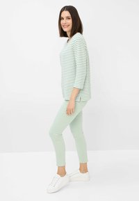 Hellgrüner gestreifter Pullover mit Dreiviertelärmeln, kombiniert mit hellgrünen Skinny-Jeans und weißen Sneakers. Schlicher Hintergrund.