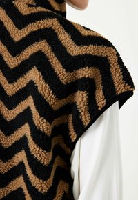 Väst med brunt och svart zick-zack-mönster av texturerad fleece, med rundad axeldesign och svarta detaljer längs kanterna.