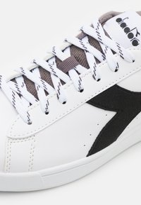 Sneaker bianco con accenti in suede nero. Presenta lacci bianchi, una linguetta in mesh e dettagli perforati. Materiale superiore sintetico liscio.