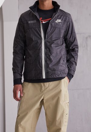 Veste coupe-vent noire avec un motif géométrique subtil, dotée d'une fermeture éclair devant et d'accents blancs, assortie à un pantalon cargo beige.